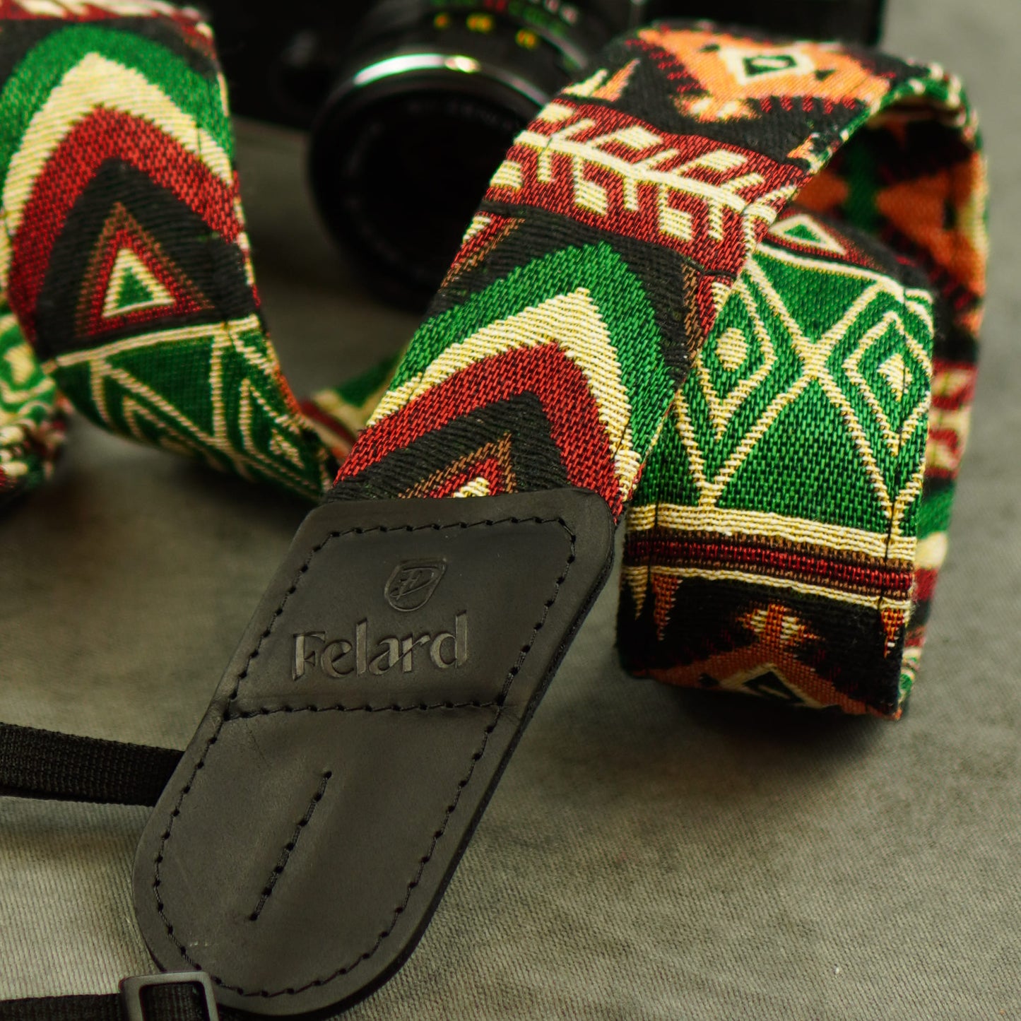 A close-up of a woven camera strap with a bold geometric pattern in green, red, black, and cream, featuring a dark gray leather brand patch that reads 'Felard'.