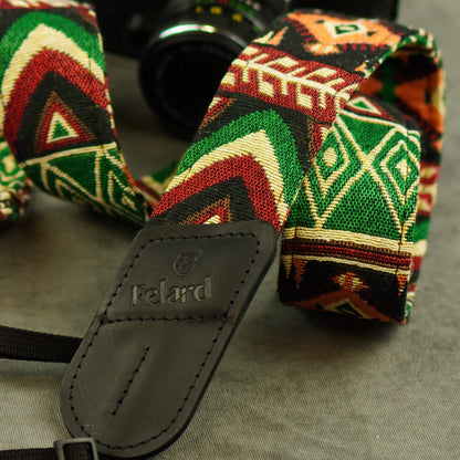 A close-up of a woven camera strap with a bold geometric pattern in green, red, black, and cream, featuring a dark gray leather brand patch that reads 'Felard'.