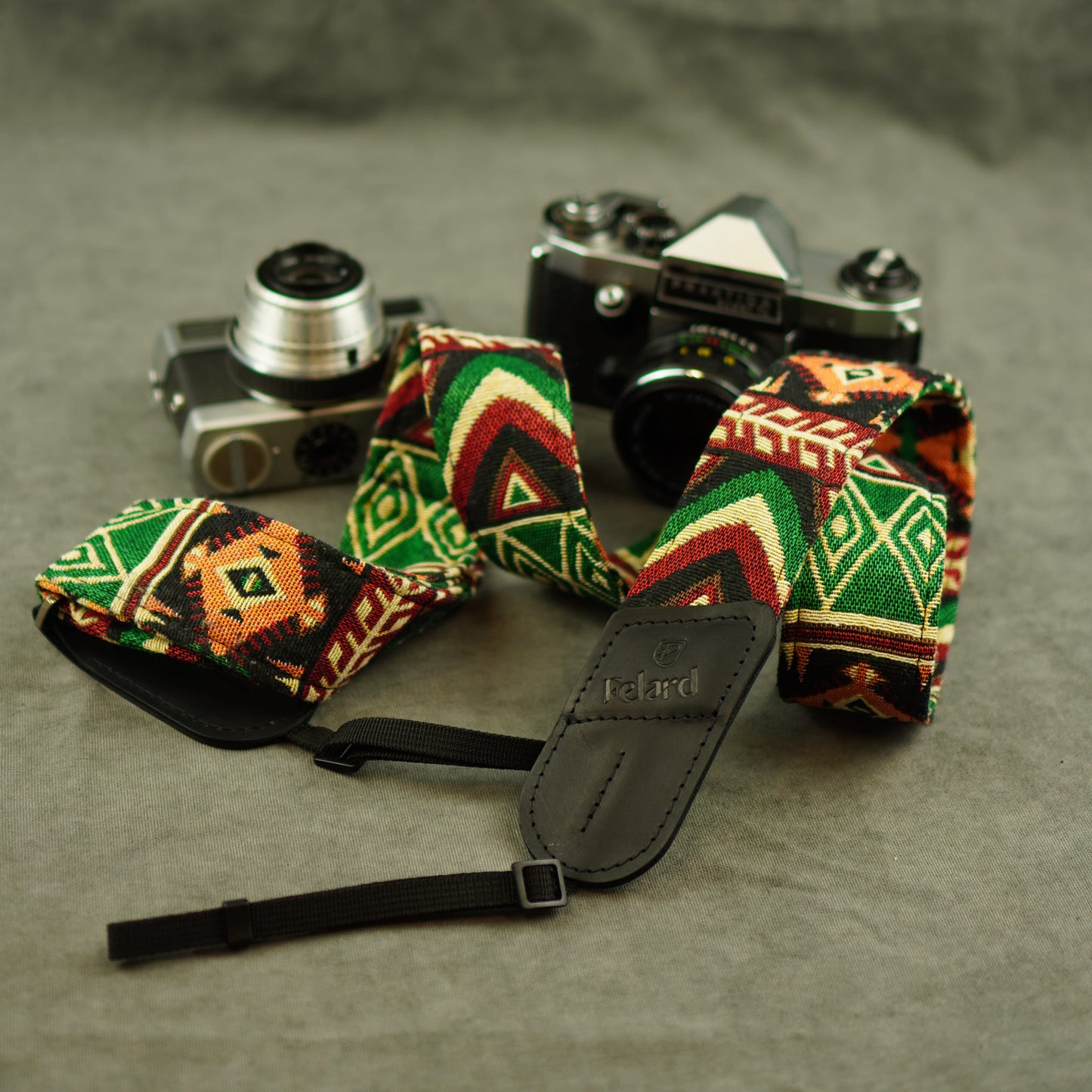 A woven camera strap with a colorful geometric pattern in green, red, orange, and black, featuring a black leather patch with the brand name 'Felard' embossed on it. The strap is displayed on a textured gray surface next to two vintage film cameras.