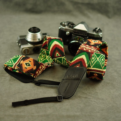A woven camera strap with a colorful geometric pattern in green, red, orange, and black, featuring a black leather patch with the brand name 'Felard' embossed on it. The strap is displayed on a textured gray surface next to two vintage film cameras.