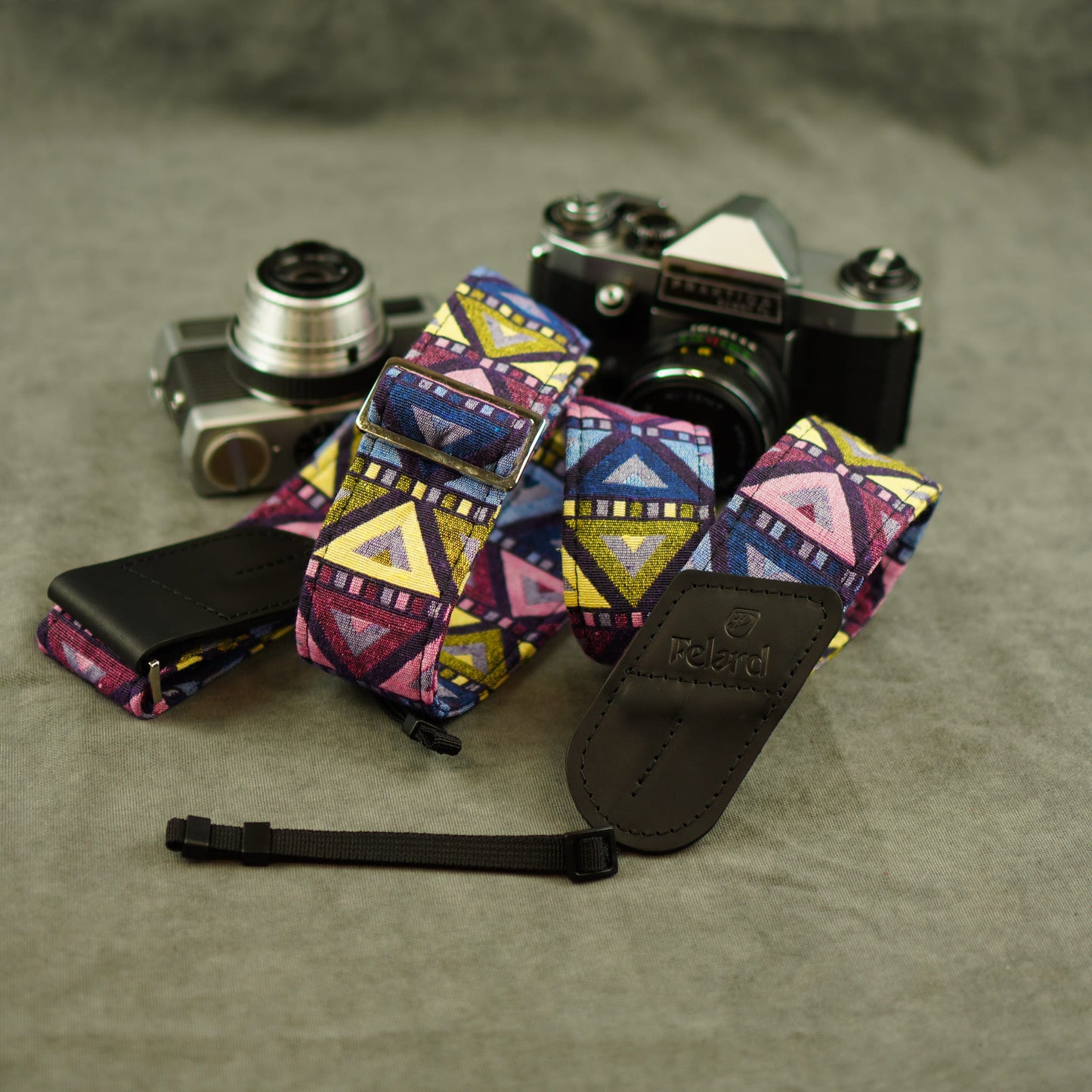 A multicolor geometric patterned camera strap with black leather ends, displayed on a textured gray surface with two cameras in the background. The brand name 'Felard' is embossed on the leather end.