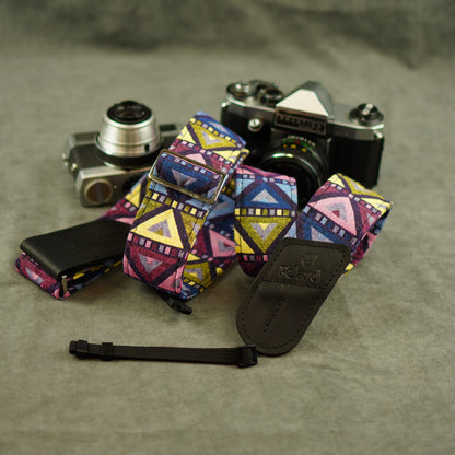 A multicolor geometric patterned camera strap with black leather ends, displayed on a textured gray surface with two cameras in the background. The brand name 'Felard' is embossed on the leather end.