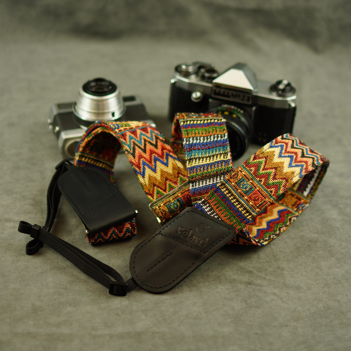 A colorful patterned camera strap with leather end displayed on top of two vintage cameras against a gray textured background.