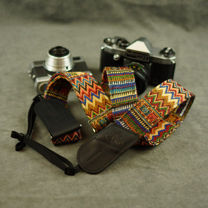 A colorful patterned camera strap with leather end displayed on top of two vintage cameras against a gray textured background.
