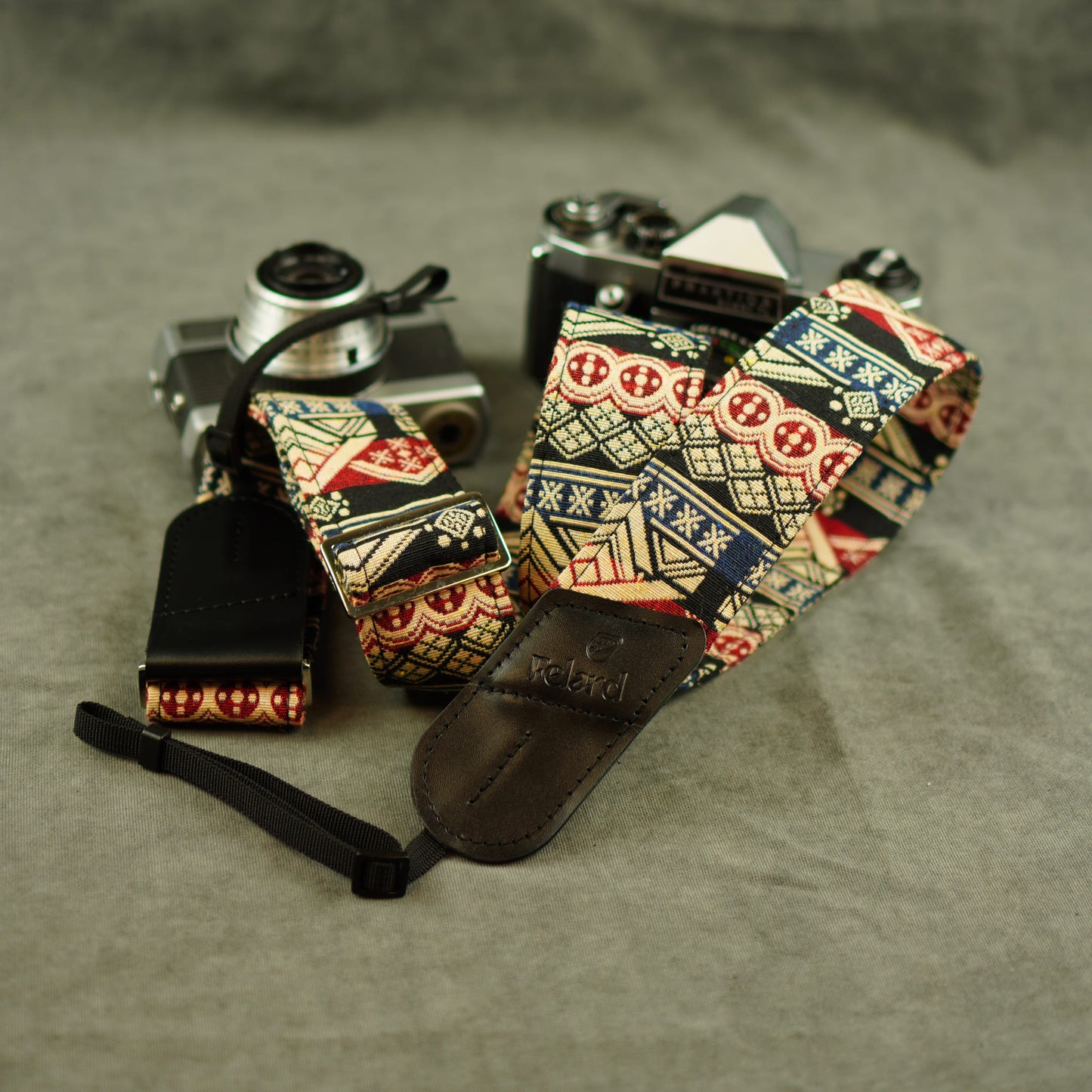 A patterned camera strap with a black leather keeper and attached wristlet, displayed on top of two vintage cameras against a gray fabric background.