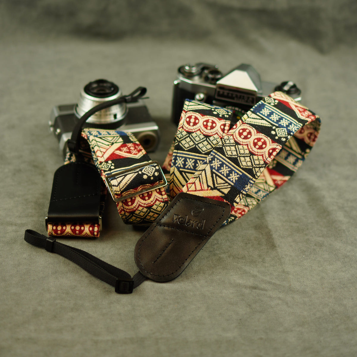 A patterned camera strap with a black leather keeper and attached wristlet, displayed on top of two vintage cameras against a gray fabric background.