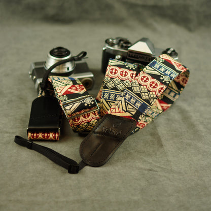 A patterned camera strap with a black leather keeper and attached wristlet, displayed on top of two vintage cameras against a gray fabric background.