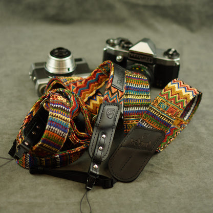 A multicolor woven camera strap with geometric patterns and a black leather keeper, displayed on top of two vintage cameras against a gray textured background.