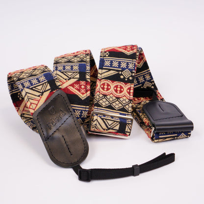A patterned camera strap with a black leather and a black adjustable webbing wristlet, displayed on a clean white background. The fabric has a complex geometric tribal pattern in red, blue, beige, and black.