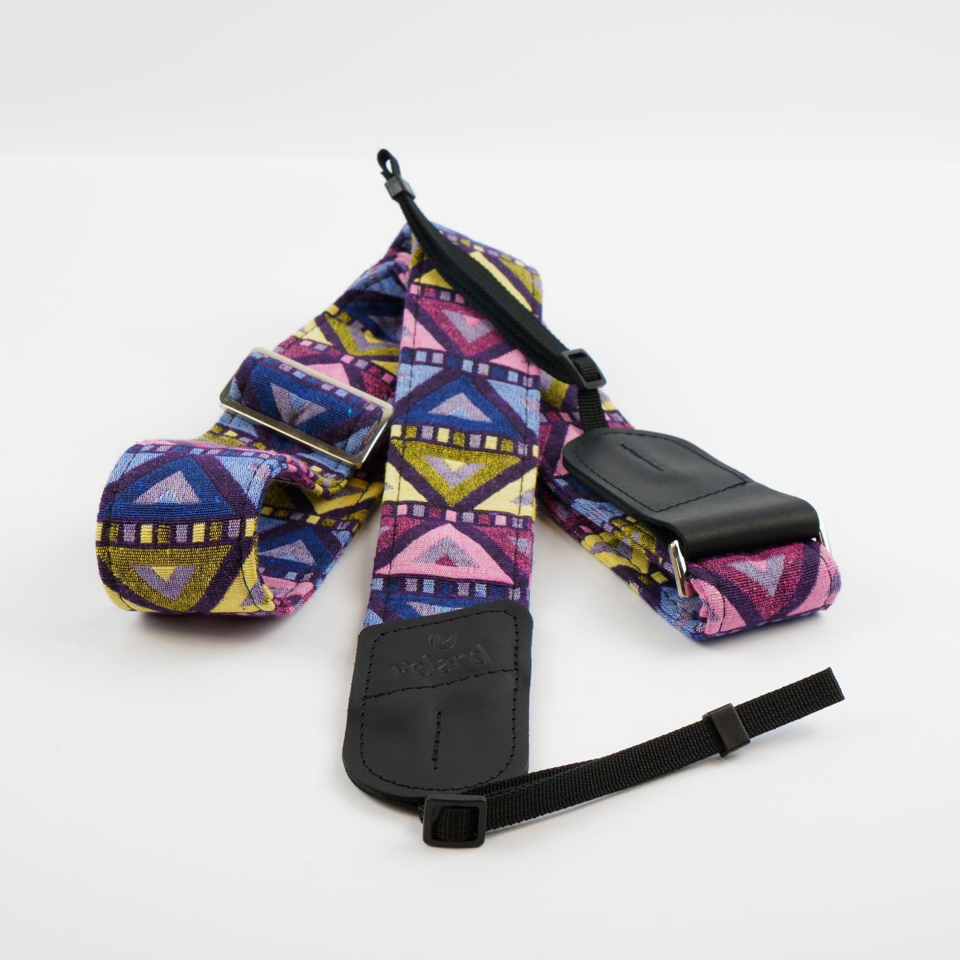 A camera strap with a colorful geometric pattern in purple, blue, yellow, and pink, featuring black leather-like buckles and a black adjustable strap.