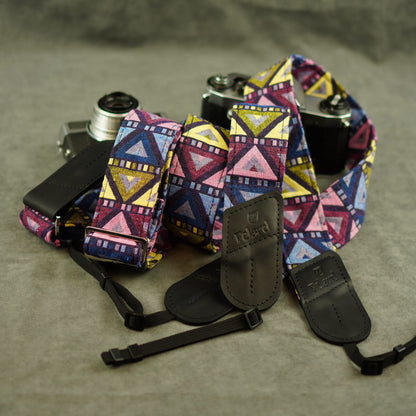 A colorful patterned camera straps with black leather attachments, laid out on a gray textured surface with two cameras partially visible behind it.