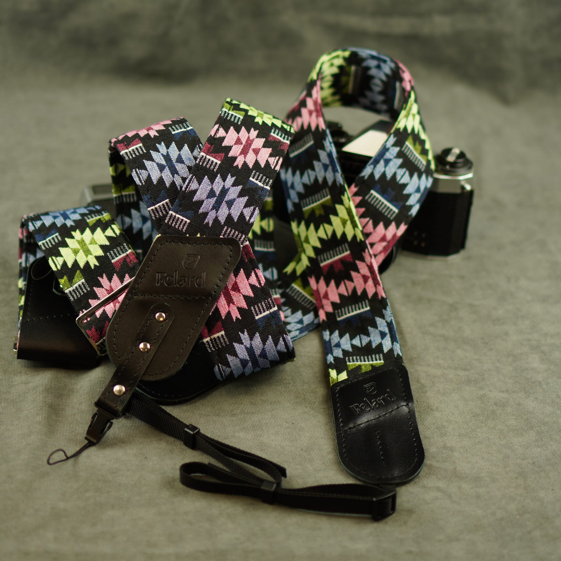A vintage-style camera strap with a colorful geometric pattern in pink, green, blue, and black, featuring dark brown leather ends and a matching camera bag, displayed on a gray textured surface.