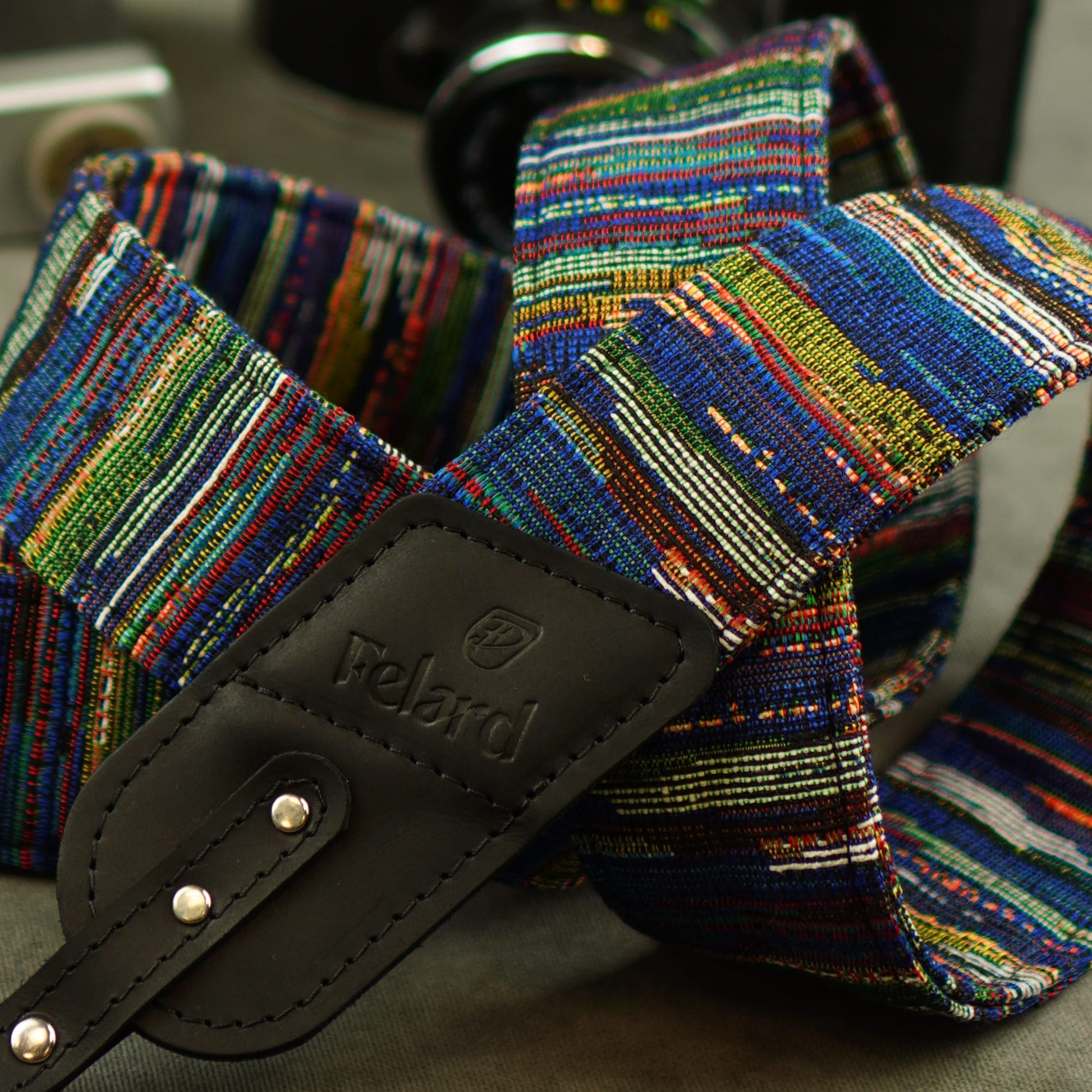 A close-up of a multicolored woven camera strap with a black leather and the brand name Felard embossed on it.