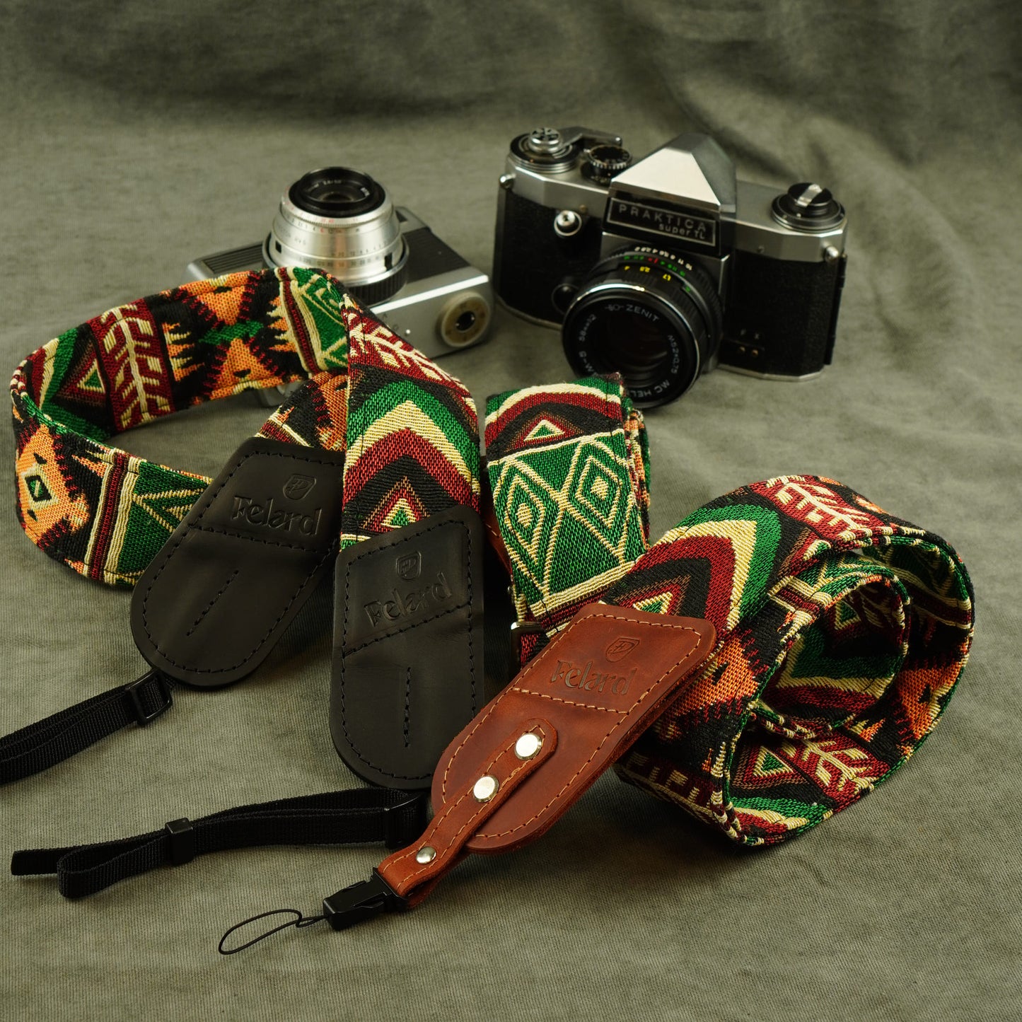 A multicolor geometric patterned camera strap with brown leather accents and black buckles, displayed on a textured gray background next to two vintage cameras.