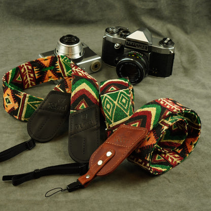 A multicolor geometric patterned camera strap with brown leather accents and black buckles, displayed on a textured gray background next to two vintage cameras.