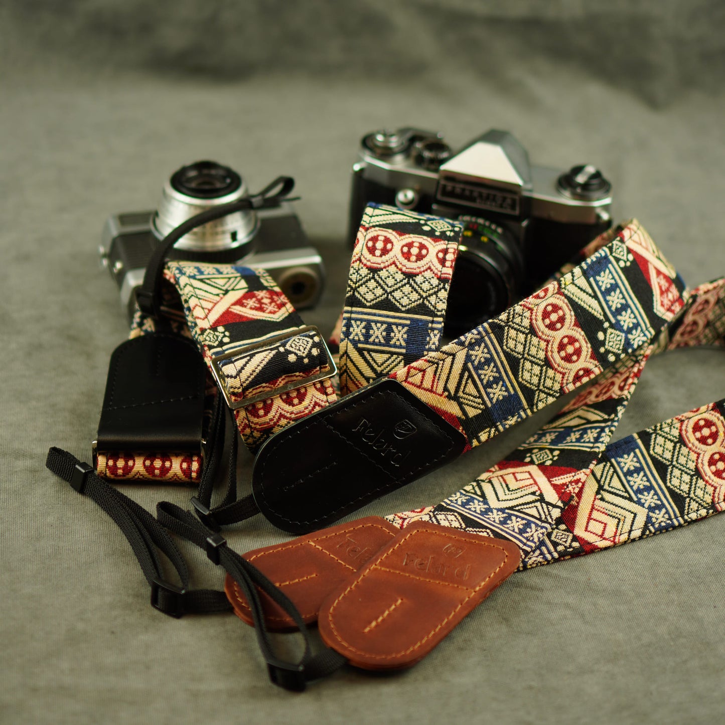 A close-up of a camera strap laid out on a gray surface, with two vintage cameras partially visible behind it. The strap has a multicolor geometric and tribal-inspired pattern in cream, navy, red, and beige, with brown leather ends and black adjustable sections.