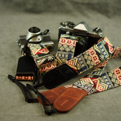 A close-up of a camera strap laid out on a gray surface, with two vintage cameras partially visible behind it. The strap has a multicolor geometric and tribal-inspired pattern in cream, navy, red, and beige, with brown leather ends and black adjustable sections.