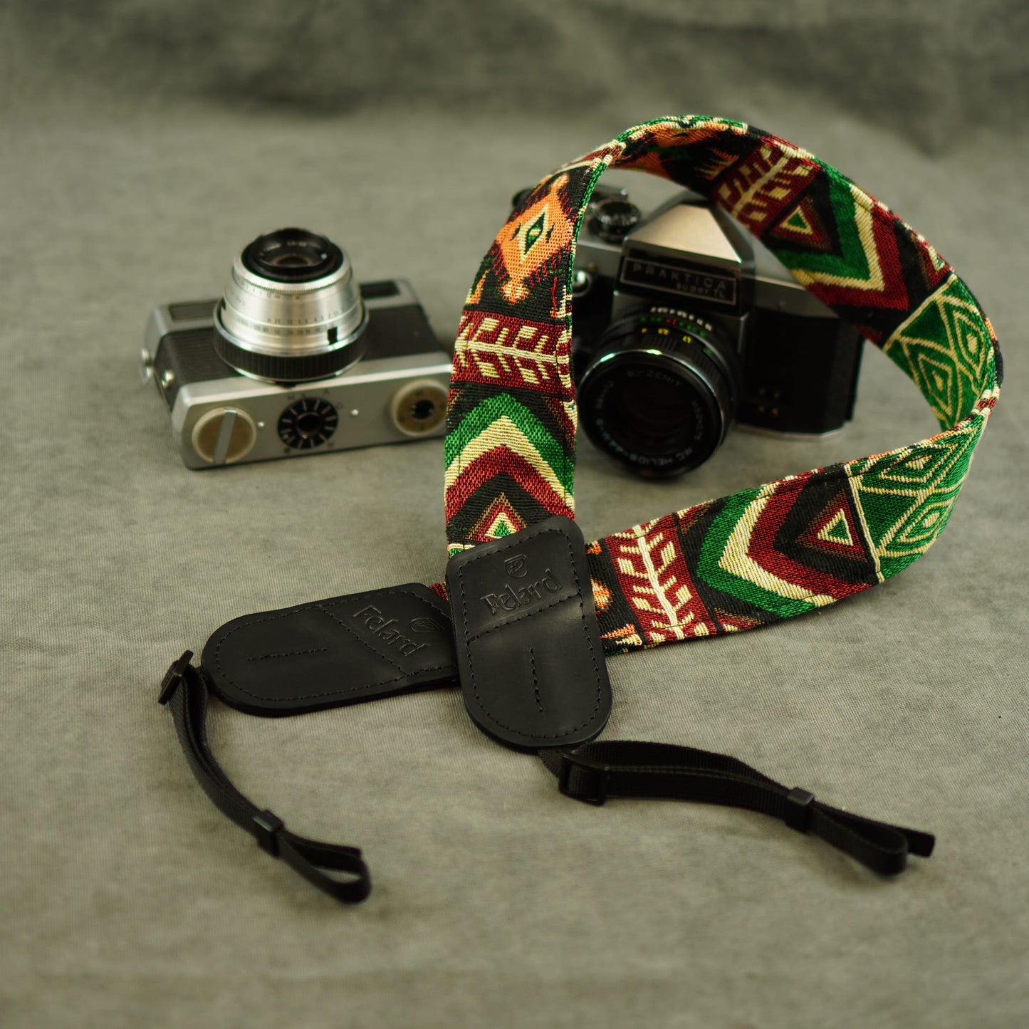 A woven camera strap with a colorful geometric pattern in green, red, yellow, and black, shown laid out on a gray textured surface next to two vintage cameras.