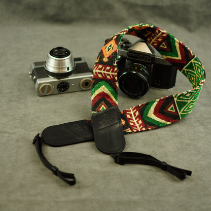 A woven camera strap with a colorful geometric pattern in green, red, yellow, and black, shown laid out on a gray textured surface next to two vintage cameras.
