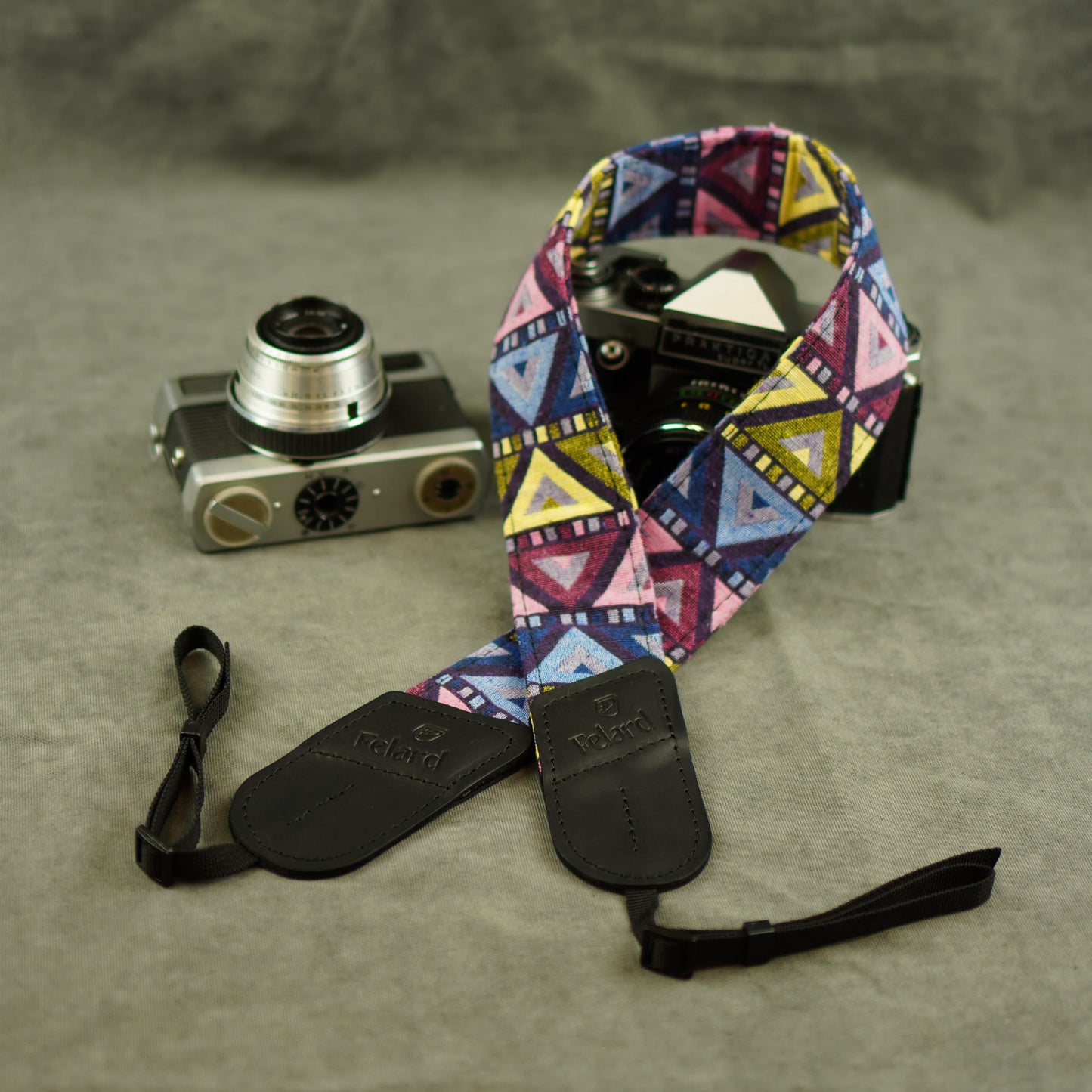 A colorful geometric patterned camera strap with black leather ends and a black wristlet, displayed on a textured gray surface next to vintage cameras.