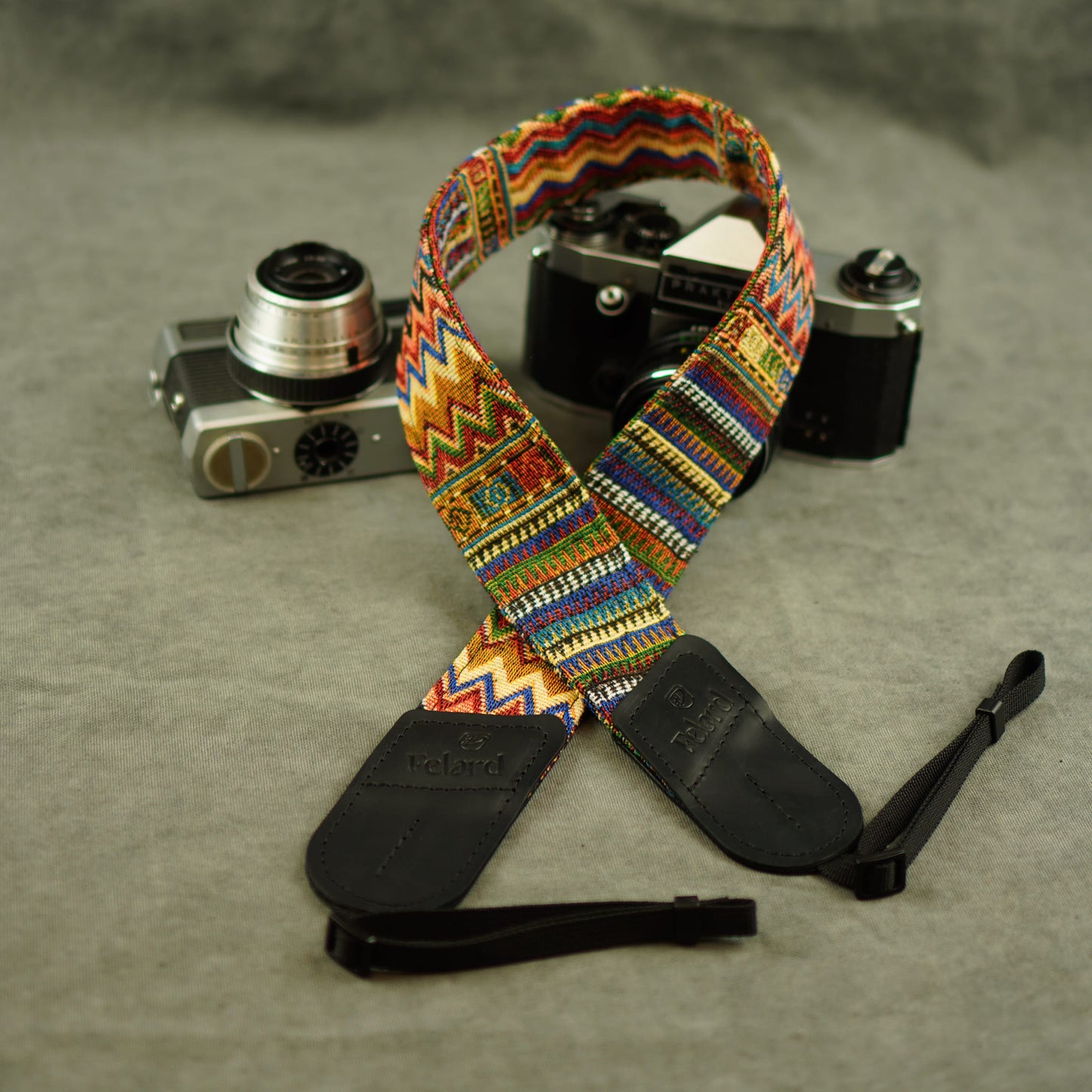 A colorful patterned camera strap with black leather ends displayed in front of two vintage film cameras on a gray textured surface.