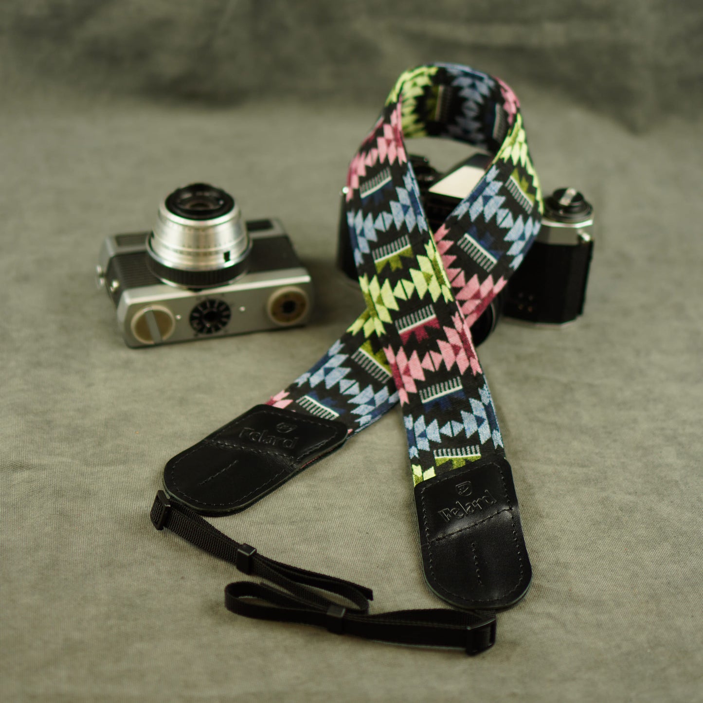 A multicolor geometric patterned camera strap with black leather ends, displayed on a textured gray surface next to two vintage cameras.