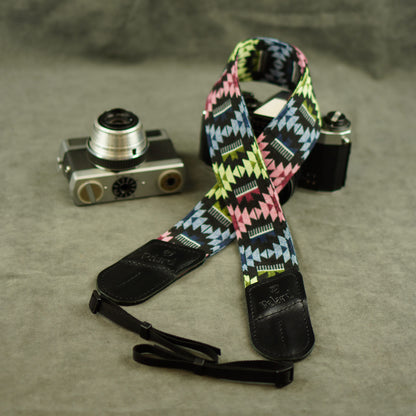 A multicolor geometric patterned camera strap with black leather ends, displayed on a textured gray surface next to two vintage cameras.