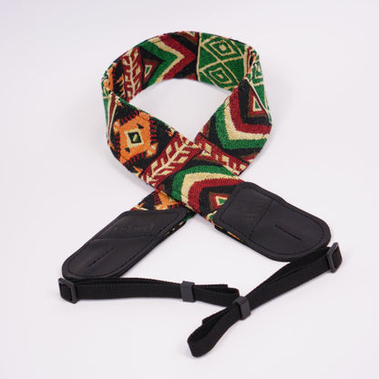 A woven camera strap featuring a vibrant Southwestern geometric pattern in green, red, orange, and beige, with black leather ends and adjustable black webbing.