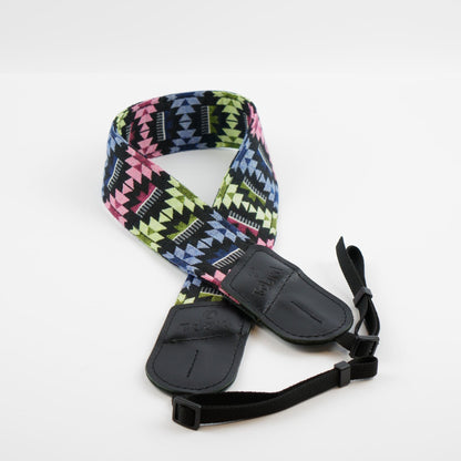 A camera strap featuring a vibrant geometric Southwestern pattern in pink, lime green, blue, and black, with black padded leather ends and an adjustable black webbing strap.