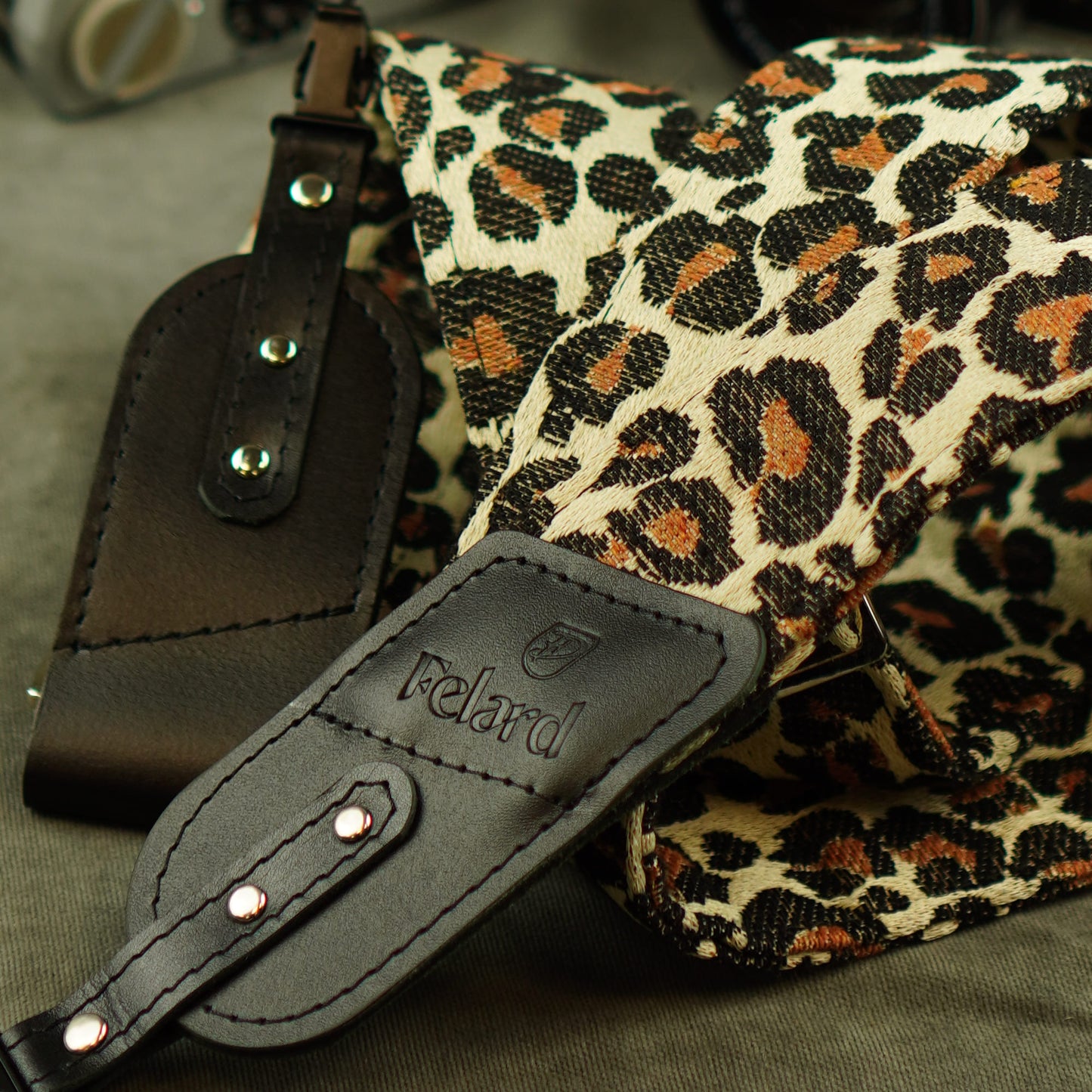 Close-up of a Felard shoulder camera strap with a leopard print canvas body and bla
ck leather attachments, including a visible Felard logo patch.
