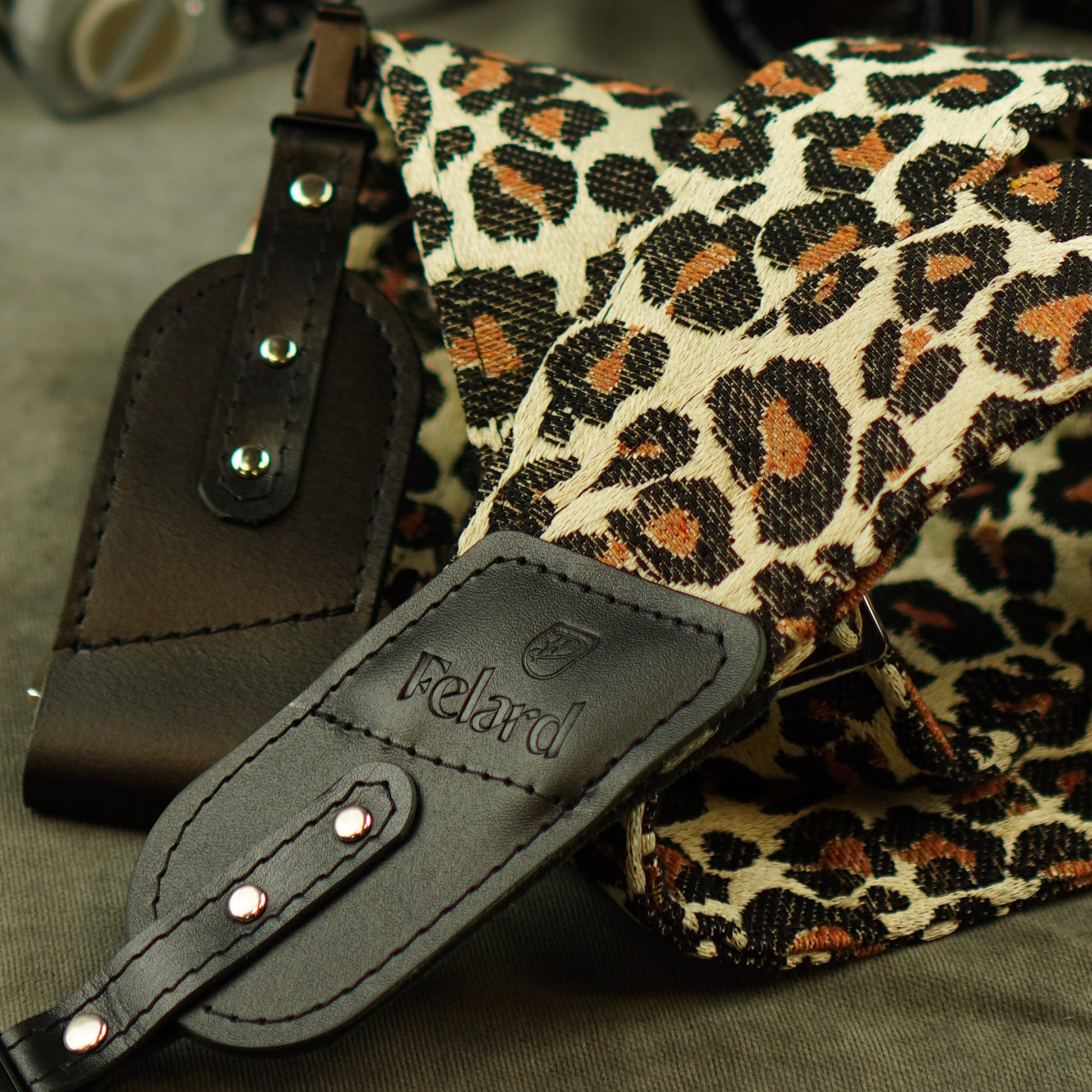 Close-up of a Felard shoulder camera strap with a leopard print canvas body and bla
ck leather attachments, including a visible Felard logo patch.