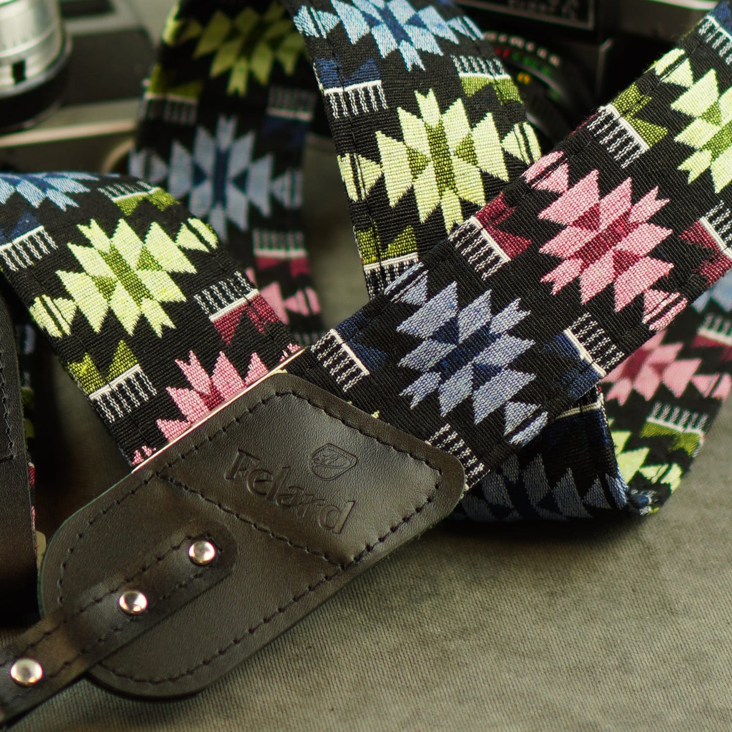 A close-up of a patterned camera strap with a black leather buckle embossed with the brand 'Felard'. The strap features a Southwestern geometric design on a black background with pink, green, blue, and white motifs.