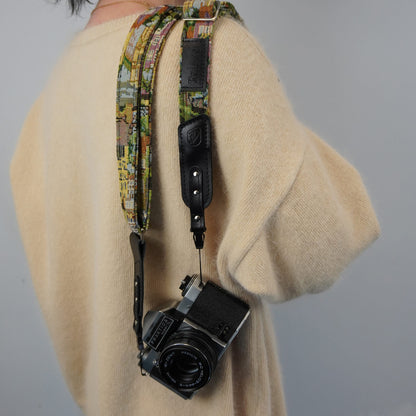 A person wearing a cream knit sweater carries a camera with a colorful patterned fabric strap and black leather accents against a plain light gray background.