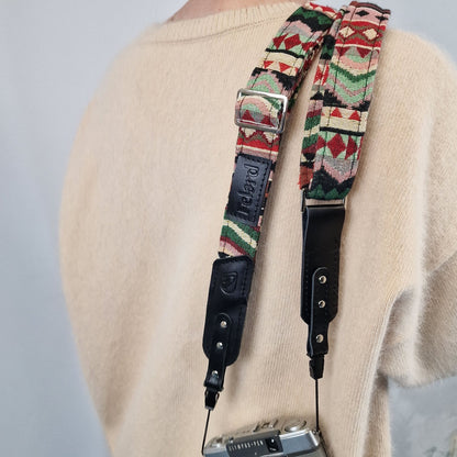 A close-up of a patterned camera strap worn over a cream knit sweater. The strap features a colorful geometric woven design in red, green, black, and white, with black leather hardware and a silver metal buckle.