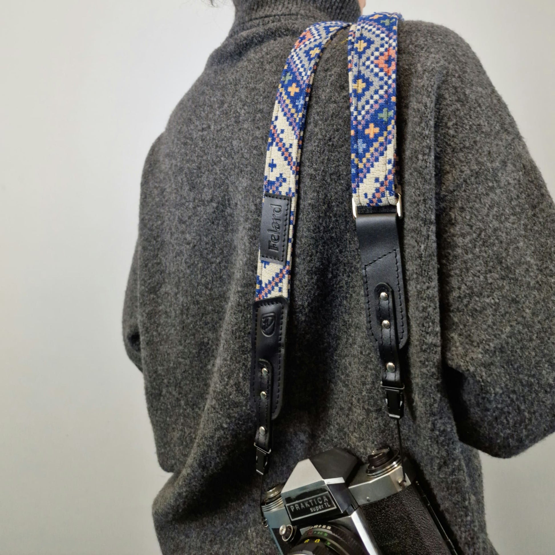 A close-up of a person from behind carrying a camera with a patterned blue, white, and orange woven strap and black leather strap details.