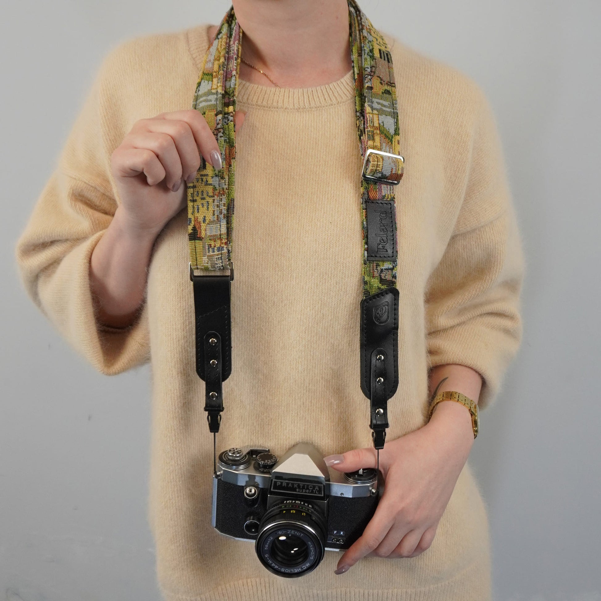 A person wearing a beige knit sweater holds a vintage-style camera with a colorful illustrated strap and black leather sections against a plain light gray background.