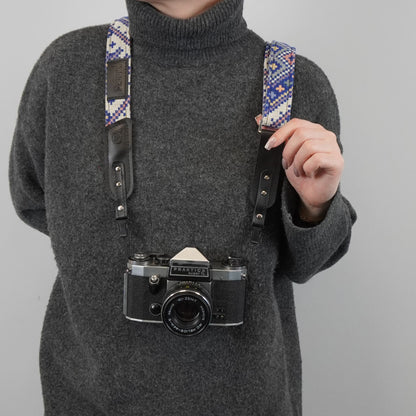 A person holds a vintage-style black camera with a patterned blue, purple, and white woven strap and black leather shoulder straps against a plain light gray background.