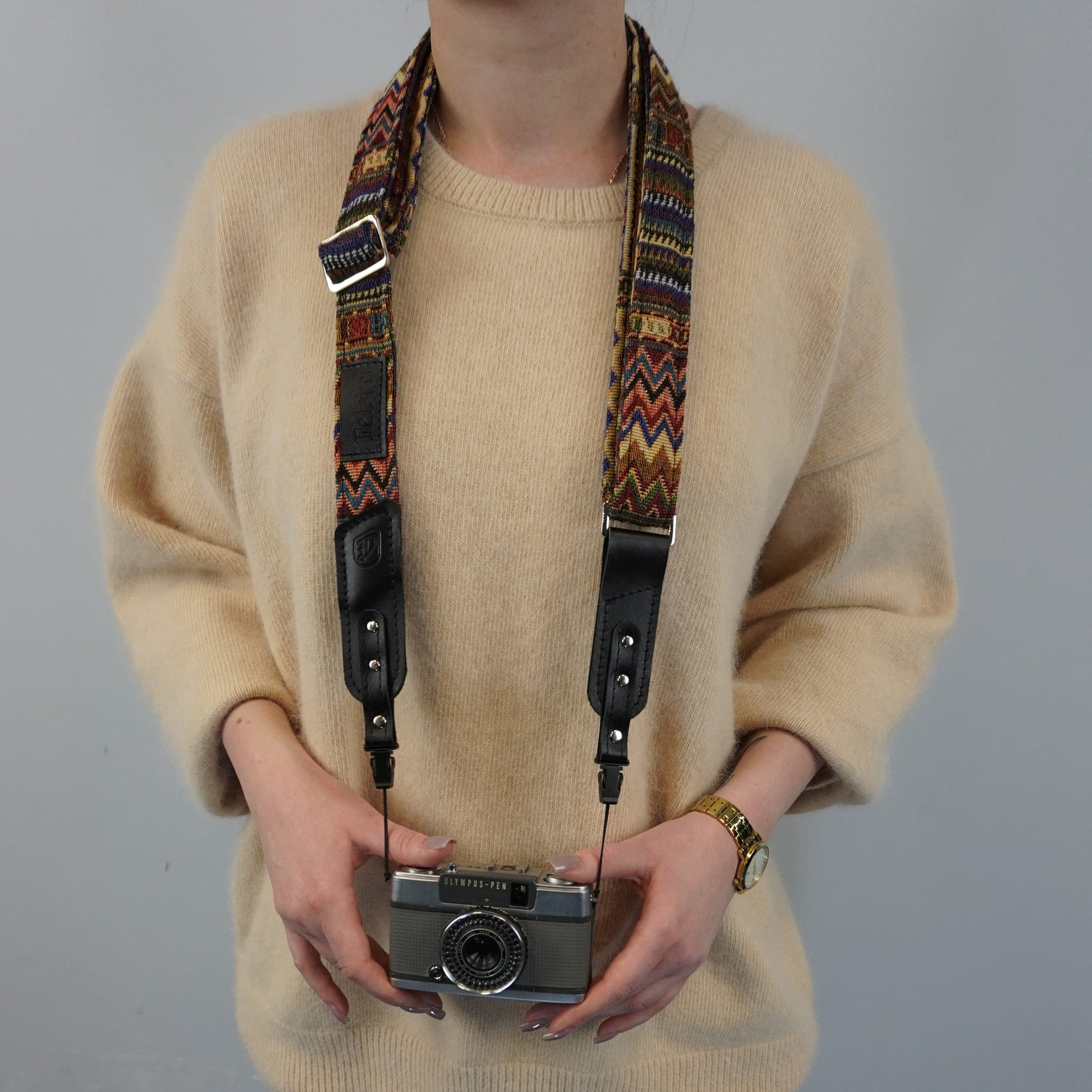A person holds a vintage silver camera with a colorful geometric patterned camera strap against a plain gray background.