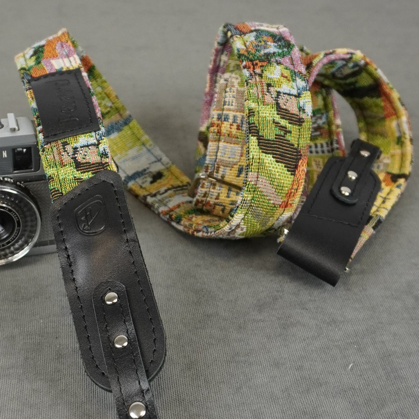 A close-up of a camera strap with a multicolor cityscape pattern and black leather accents, shown next to a vintage camera on a gray textured background.