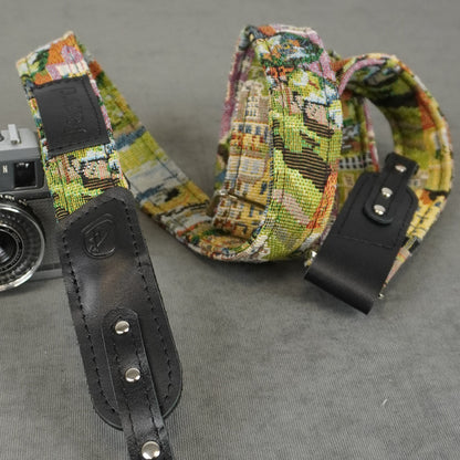 A close-up of a camera strap with a multicolor cityscape pattern and black leather accents, shown next to a vintage camera on a gray textured background.