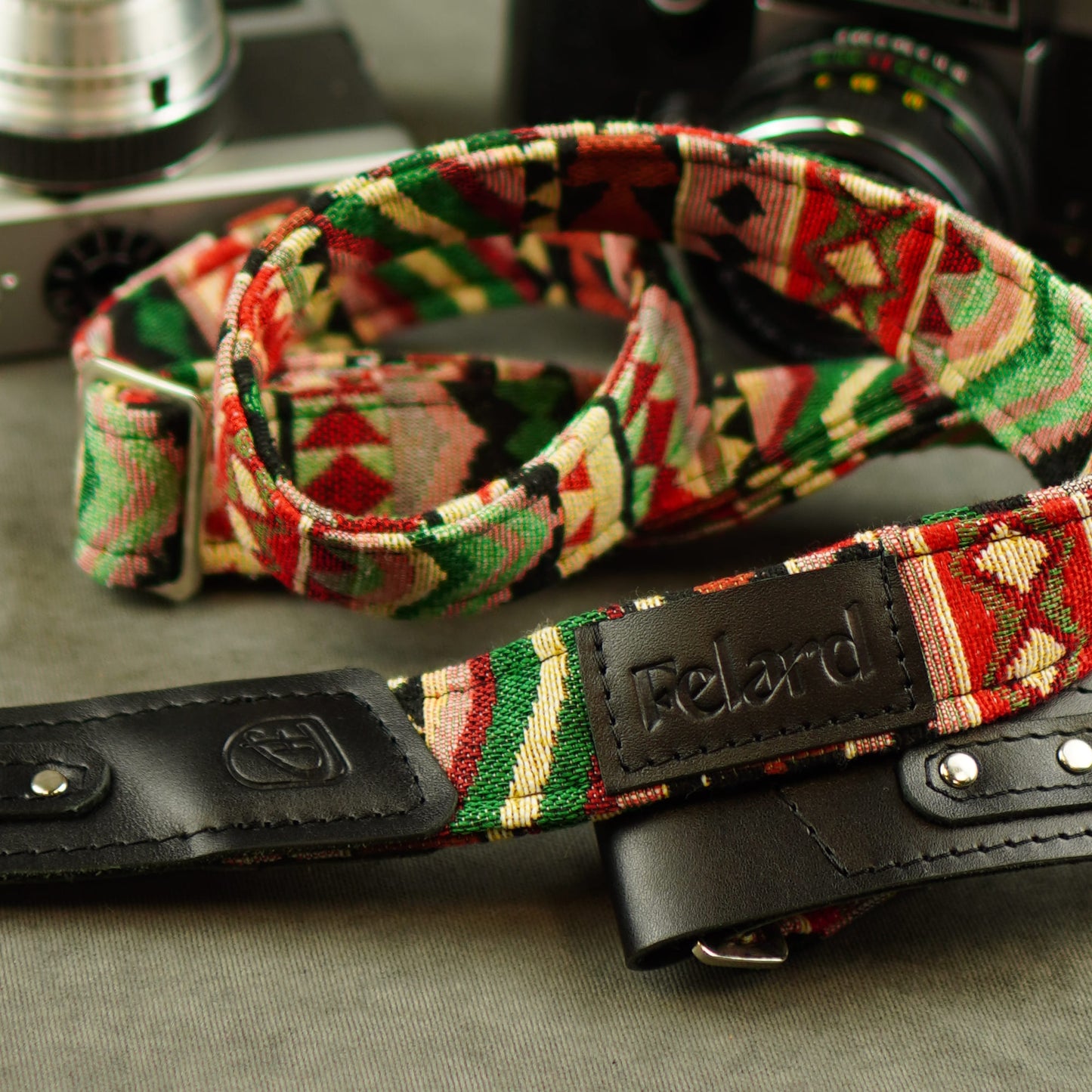 A close-up of a camera strap with a colorful geometric patterned fabric and black leather accents, featuring a leather patch engraved with the brand name 'Felard'.