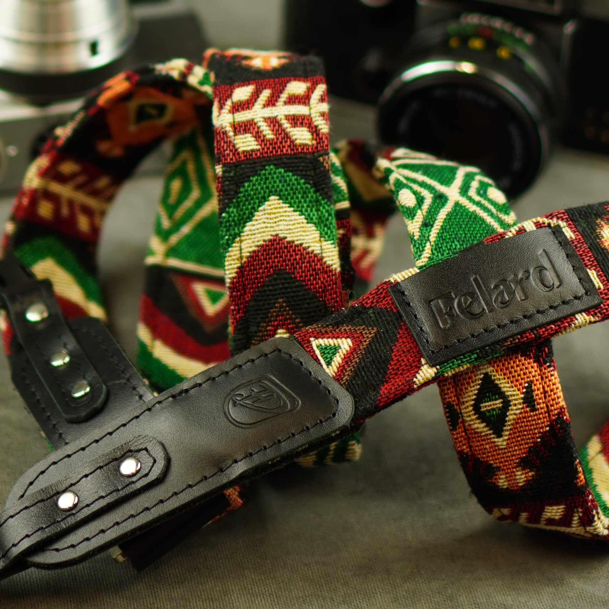 A close-up of a Felard camera strap featuring a colorful geometric woven pattern in green, red, orange, and cream, with black leather hardware.