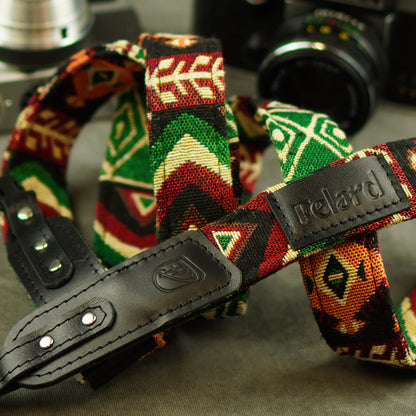 A close-up of a Felard camera strap featuring a colorful geometric woven pattern in green, red, orange, and cream, with black leather hardware.