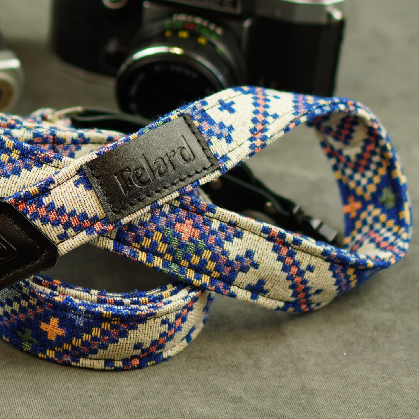 A close-up of a woven camera strap with a geometric pattern in blue, orange, yellow, and white, featuring a black leather brand patch that reads 'Felard'. A vintage camera is blurred in the background on a gray surface.