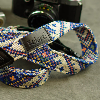 A close-up of a woven camera strap with a geometric pattern in blue, orange, yellow, and white, featuring a black leather brand patch that reads 'Felard'. A vintage camera is blurred in the background on a gray surface.