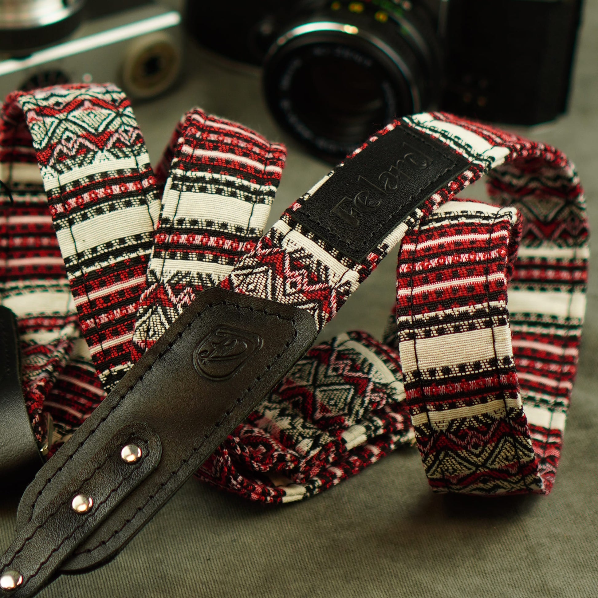 A camera strap with a red, black, and cream geometric pattern, featuring a black leathe and a black leather brand patch that reads 'Felard'.
