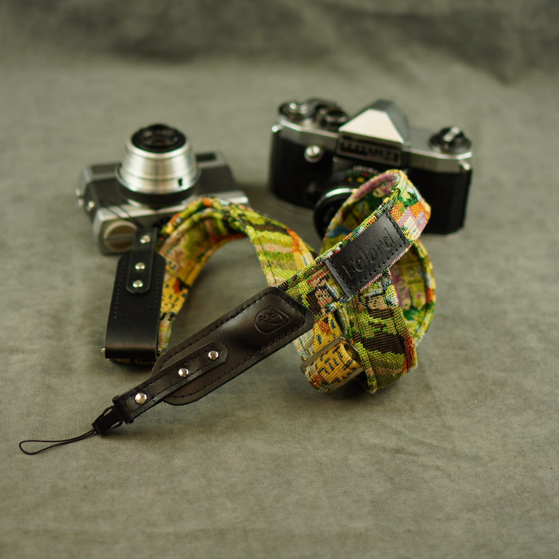 A colorful patterned camera strap with black leather accents and silver metal rivets, displayed on a textured gray surface with two vintage cameras in the background.