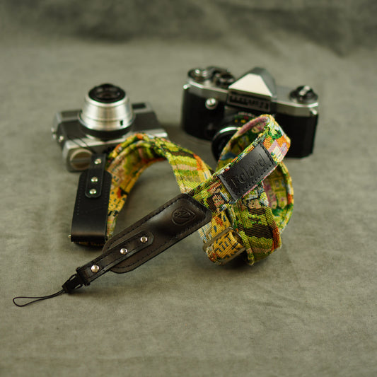 A colorful patterned camera strap with black leather accents and silver metal rivets, displayed on a textured gray surface with two vintage cameras in the background.
