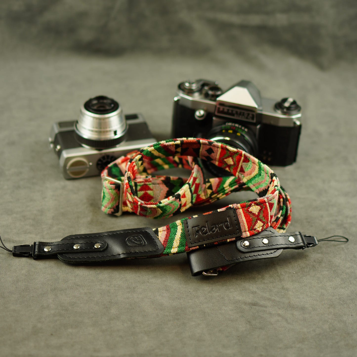 A camera strap with a colorful geometric pattern and a black leather section, displayed in front of two vintage cameras on a textured gray surface.