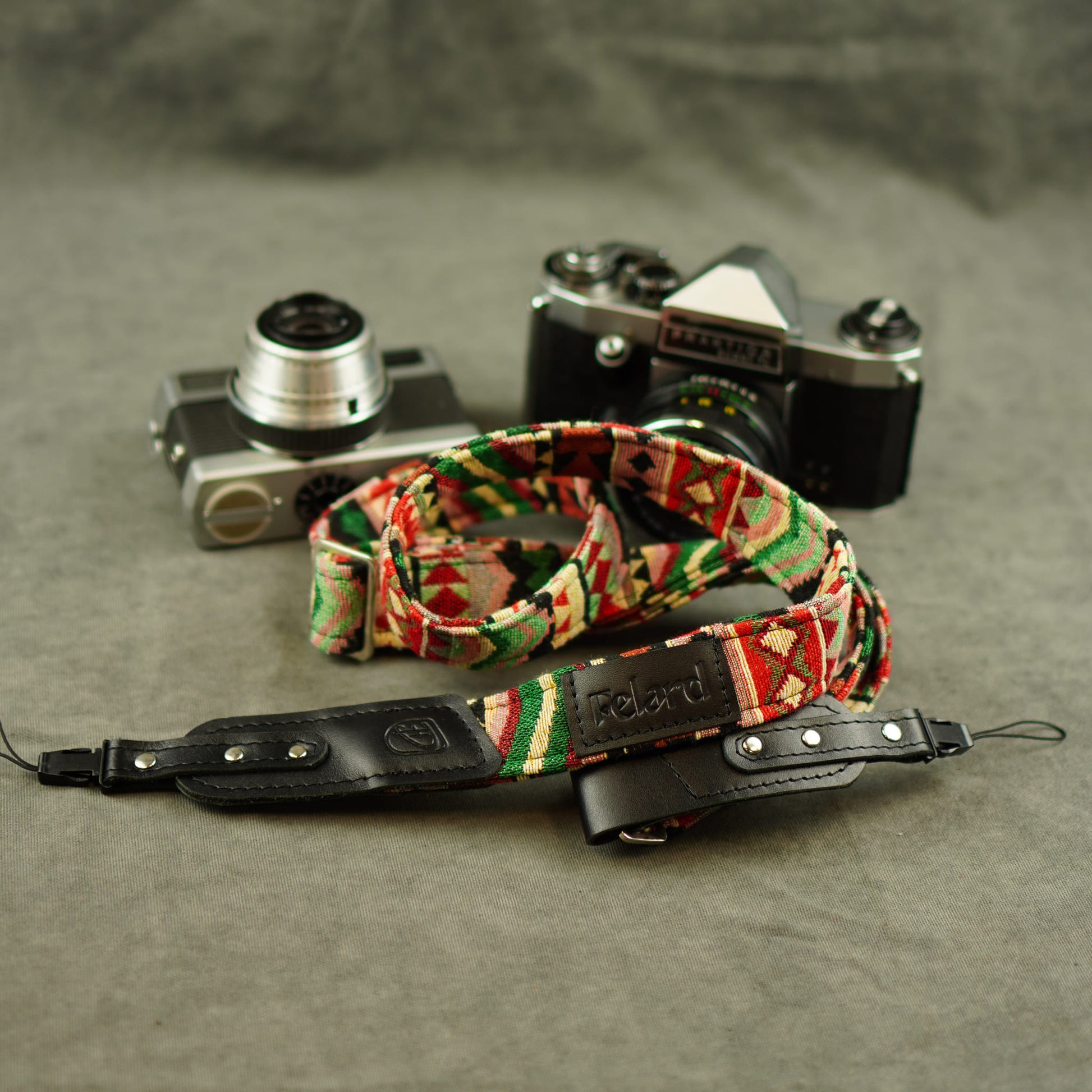 A camera strap with a colorful geometric pattern and a black leather section, displayed in front of two vintage cameras on a textured gray surface.